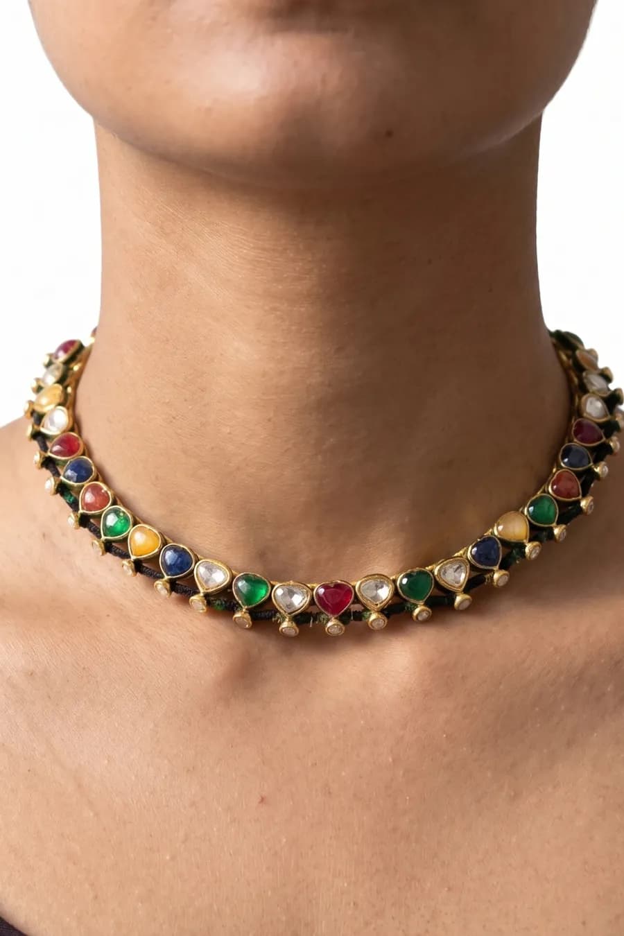 A gold navratna hasli worn at the collarbone — heart-cut ruby, emerald, sapphire and topaz with kundan between them, strung on a dark cord.